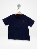 Front and back view of a Ralph Lauren baby boys' navy blue short-sleeved t-shirt. The front features a small, signature red embroidered polo player logo on the left chest. The shirt is displayed on a wooden hanger against a colorful Tiny Thrift Store backdrop. A close-up shows the authentic Ralph Lauren neck label indicating size 18M.