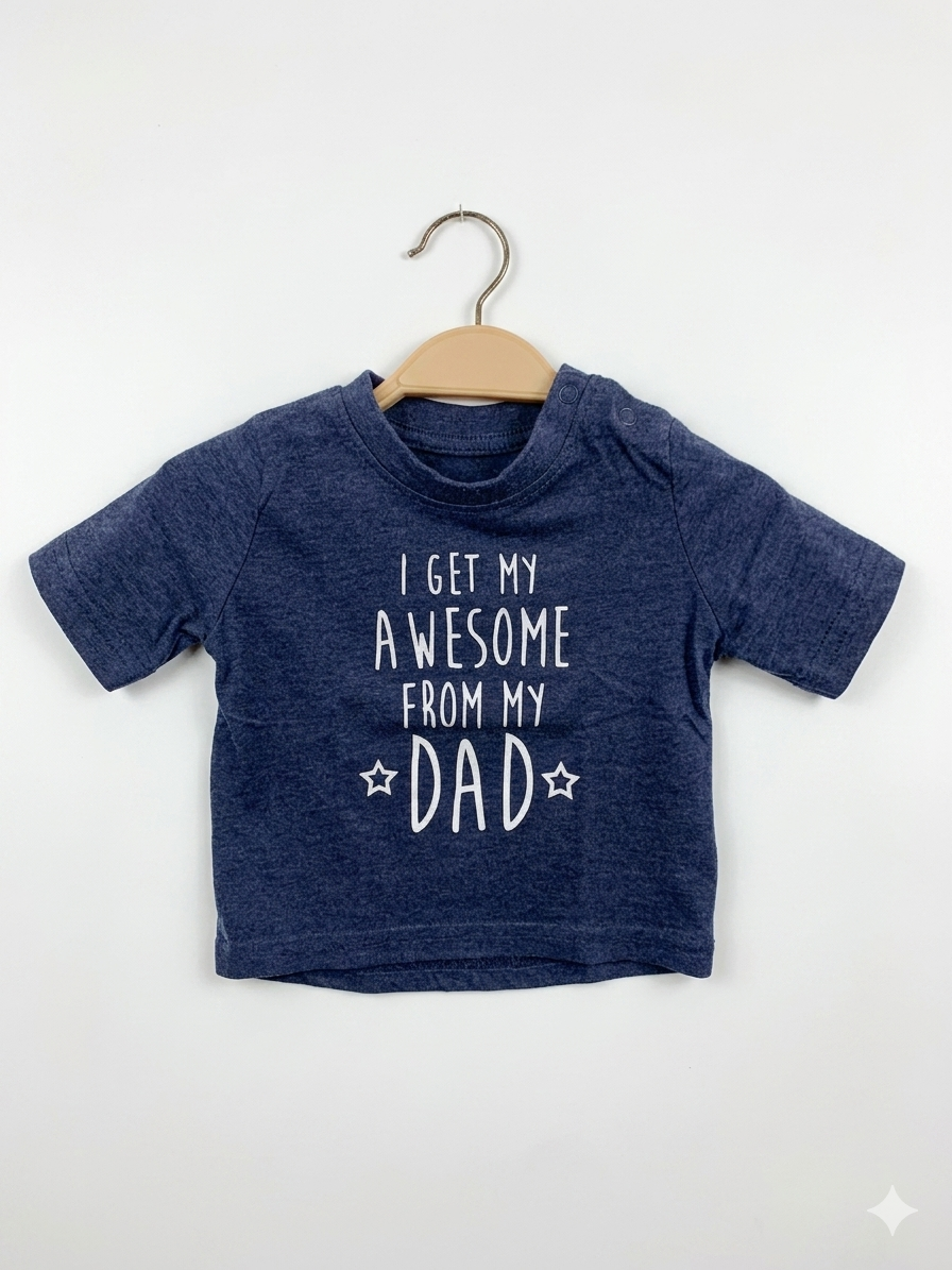 Front and back view of a small heathered navy blue infant t-shirt by Primark hanging on a wooden hanger. It features a bold white text print on the chest reading: "I Get My Awesome From My ☆ DAD ☆" and contrasting black buttons on the shoulder placket. Close-up shows a white size stamp label reading "SIZE 3–6 Months / 68CM" and "MADE IN INDIA". The backdrop is a colorful thrift store checkered wall.