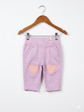 A pair of light pink baby trousers by Lindex featuring two peach-colored heart patches on the knees. The pants are hanging on a white clip hanger against a colorful Tiny Thrift Store backdrop.