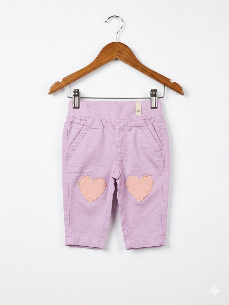 A pair of light pink baby trousers by Lindex featuring two peach-colored heart patches on the knees. The pants are hanging on a white clip hanger against a colorful Tiny Thrift Store backdrop.
