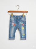 A pair of Next brand baby girl denim jeans featuring various embroidered patches like a bunny, cat, and sun on the front. The back view shows a purple butterfly patch on the pocket. The jeans are displayed on a white hanger against the Tiny Thrift Store backdrop.