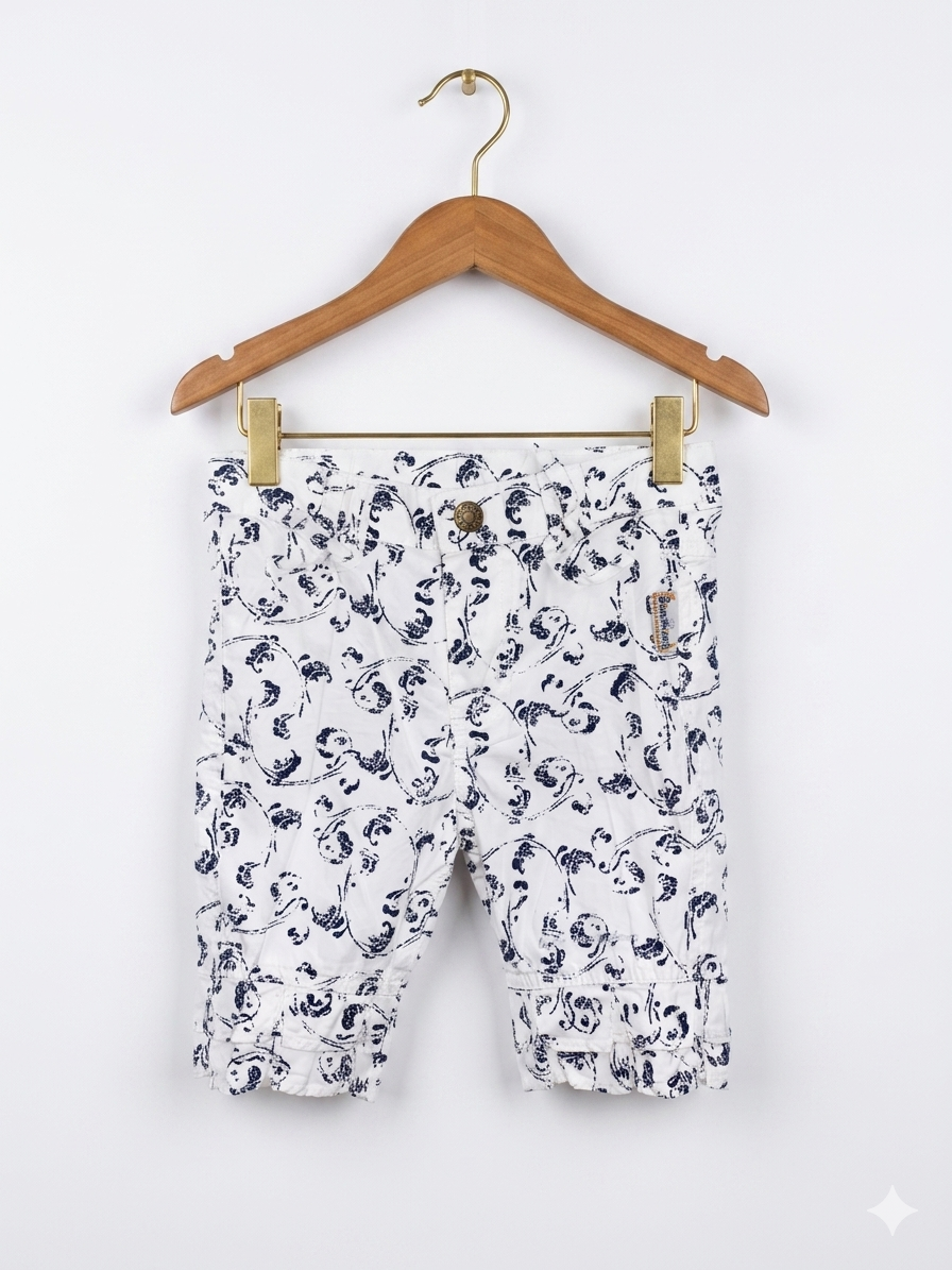 A pair of white girls' capri pants with a navy blue floral print hanging on a white hanger against a colorful Tiny Thrift Store backdrop. The pants feature ruffle hems and small bows at the waist.