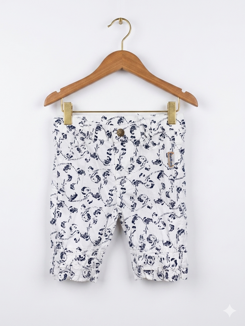 A pair of white girls' capri pants with a navy blue floral print hanging on a white hanger against a colorful Tiny Thrift Store backdrop. The pants feature ruffle hems and small bows at the waist.