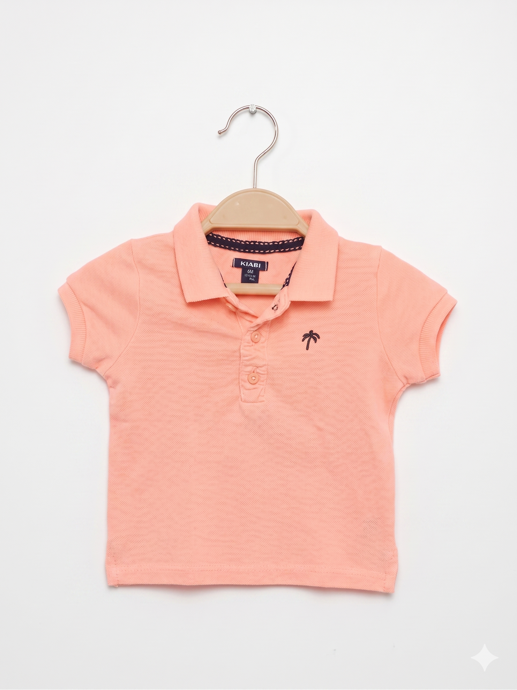 Front and back view of a peach-colored Kiabi baby polo shirt. It features a collar, three buttons, and a small black palm tree embroidered on the chest.