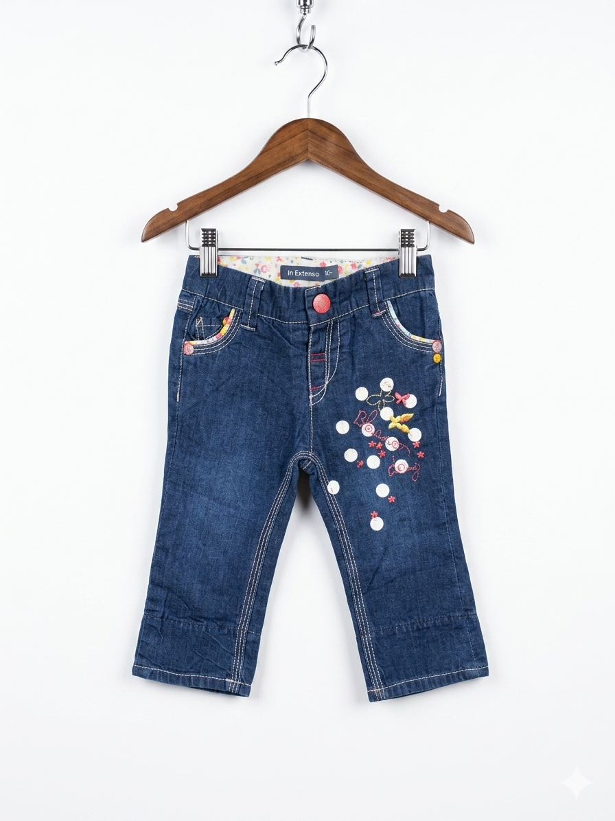 A pair of In extenso brand baby girl's dark blue denim jeans, size 62cm, are hanging from a hanger against a colorful background. The jeans feature white polka dots and floral embroidery on the left leg, colorful floral fabric pocket linings, and an elasticated waist. The label with the brand name "In extenso" and size "62cm" is visible in one close-up shot.