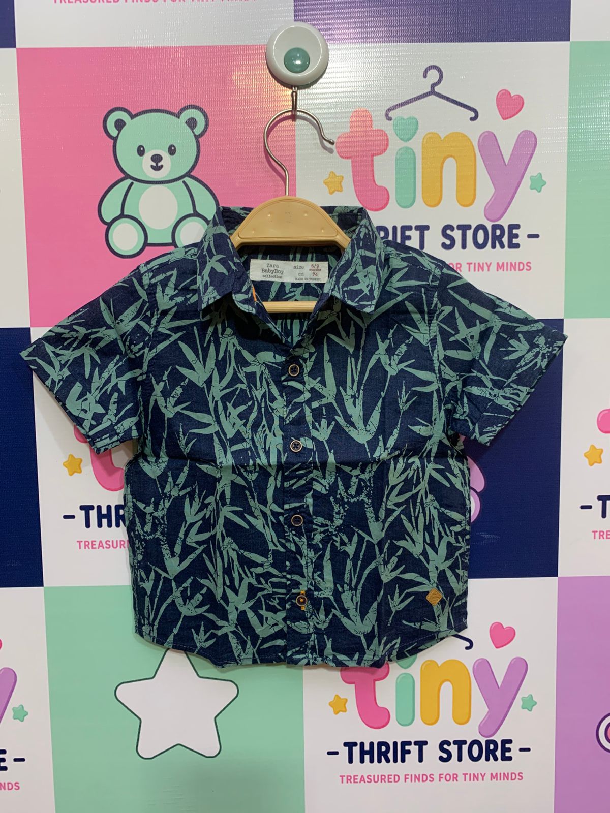 Front and back view of a ZARA Baby Boy short-sleeved button-down shirt hanging against a patterned backdrop. The shirt is navy blue with a detailed light green bamboo leaf pattern. It has a collar and brown buttons down the front. A close-up shows the white fabric brand tag, which reads "Zara BabyBoy collection," "size 6/9 months," "cm 74," and "MADE IN TURKEY," all set against the same fabric pattern.