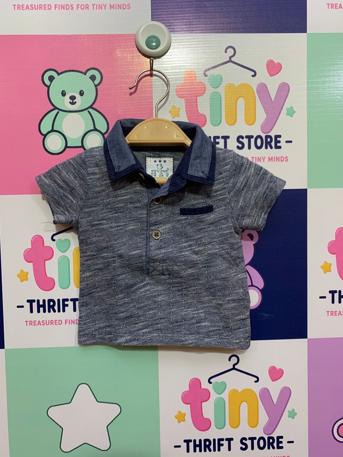Front and back view of a textured navy blue baby boy's short-sleeve polo shirt. It has white polka dots, a contrast blue denim collar, three buttons, and a small pocket. Close-up shows the white and star-patterned "Early Days" tag, reading 'Made in India'.