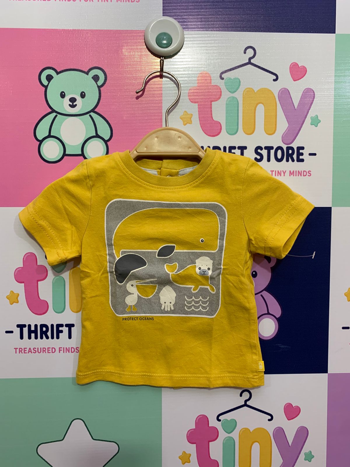 Front and back view of a small mustard yellow infant t-shirt by Obaïbi hanging against a checkered background. The front has a grey marine-life print and text. The back shows a column of yellow snap buttons from neck to hem. Close-up shows the internal label with the "Obaïbi" logo, '100% cotton', '3m / 59cm', and 'Designed in France'.