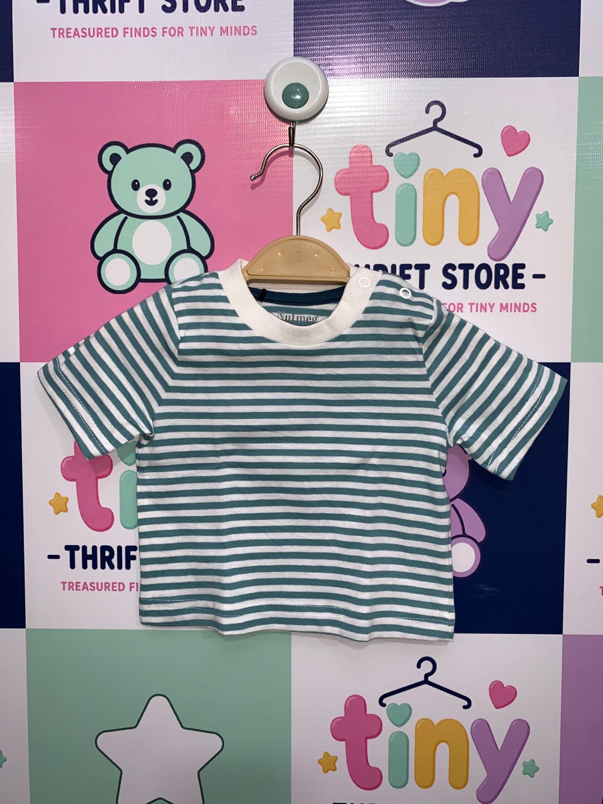 Front and back view of a teal and white striped baby t-shirt hanging against a colorful thrift store backdrop. Close-up shows the brand label "Nutmeg" with size details "6-9 months, up to 9.5kg, 74cm" and "100% cotton" material information.