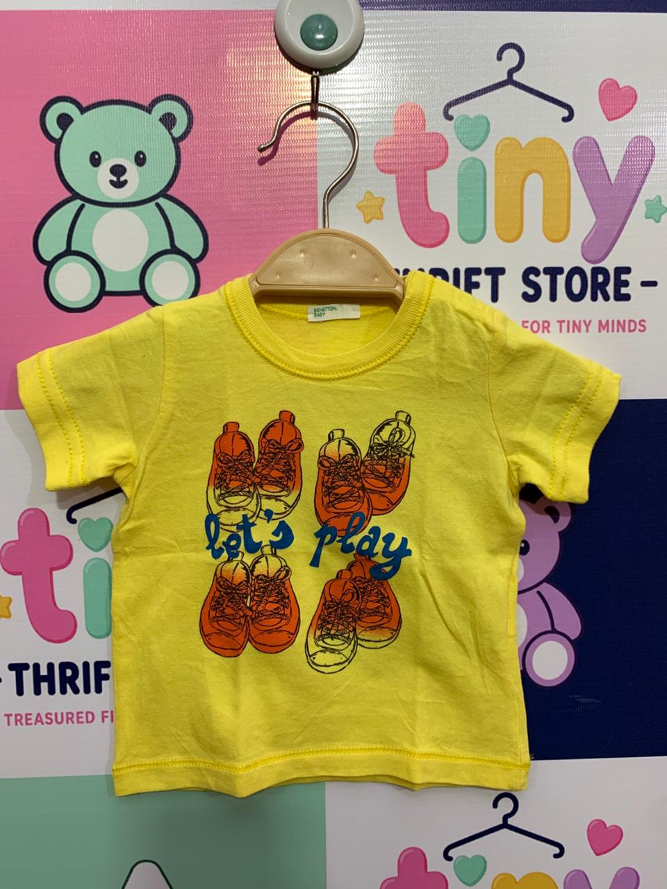 Front and back view of a small bright yellow infant t-shirt by United Colors of Benetton hanging on a hanger. The front has a graphic of four sneakers with the text "Let's play". Close-up shows the brand label "BENETTON BABY" and the size/care label reading "SIZE 56" and "1-3 MONTHS". The backdrop is a colorful thrift store checkered wall.