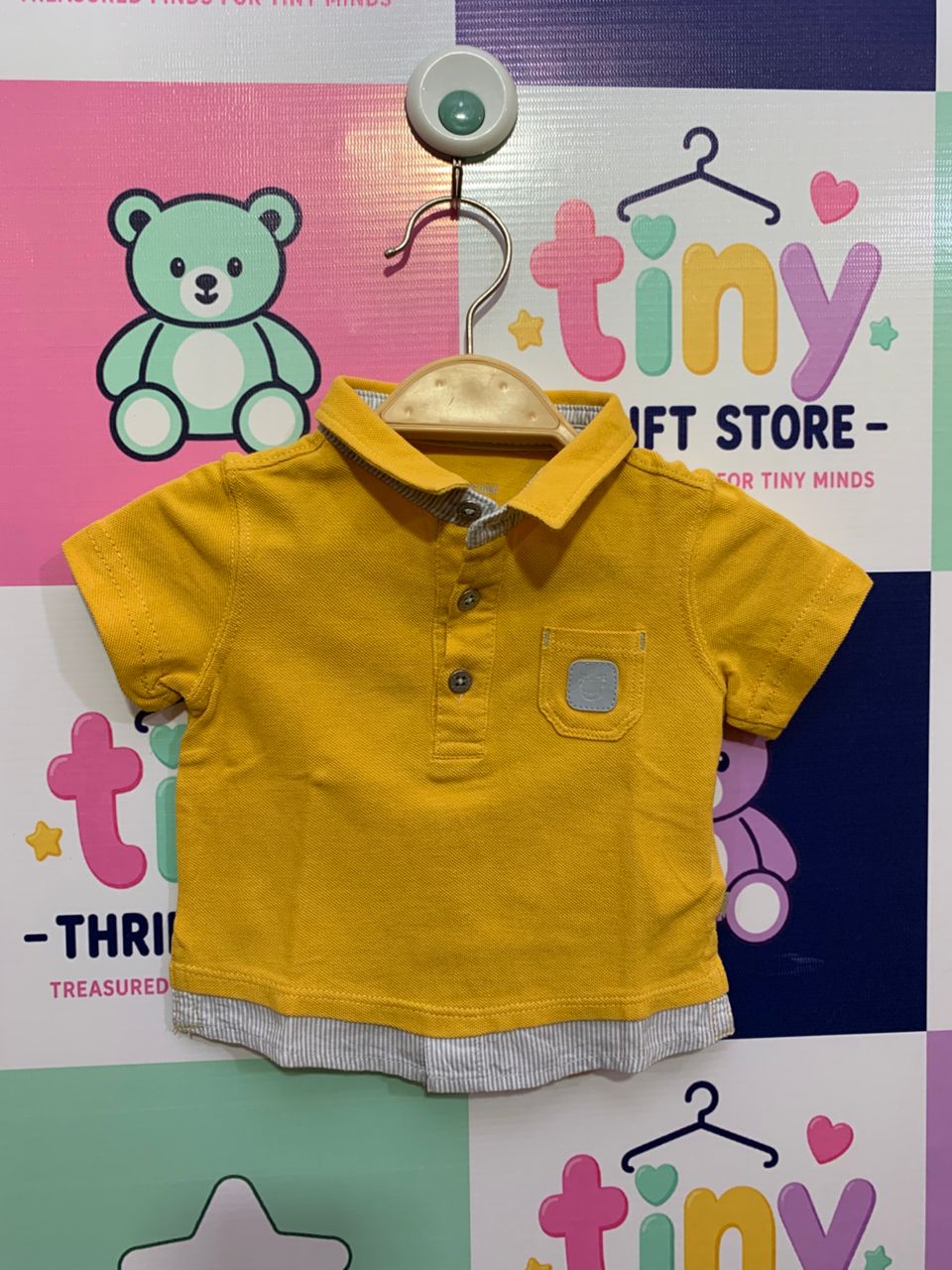 Front and back view of a small mustard-yellow infant t-shirt by OBAÏBI hanging on a wooden hanger. It features light blue striped accents on the collar and bottom hem with a small bear patch on the chest pocket. Inside the collar, a silver stamp label reads "Designed in France" and "Size 3m/59cm". The backdrop is a colorful checkered thrift store wall.