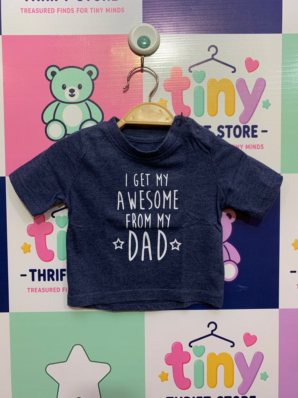 Front and back view of a small heathered navy blue infant t-shirt by Primark hanging on a wooden hanger. It features a bold white text print on the chest reading: "I Get My Awesome From My ☆ DAD ☆" and contrasting black buttons on the shoulder placket. Close-up shows a white size stamp label reading "SIZE 3–6 Months / 68CM" and "MADE IN INDIA". The backdrop is a colorful thrift store checkered wall.