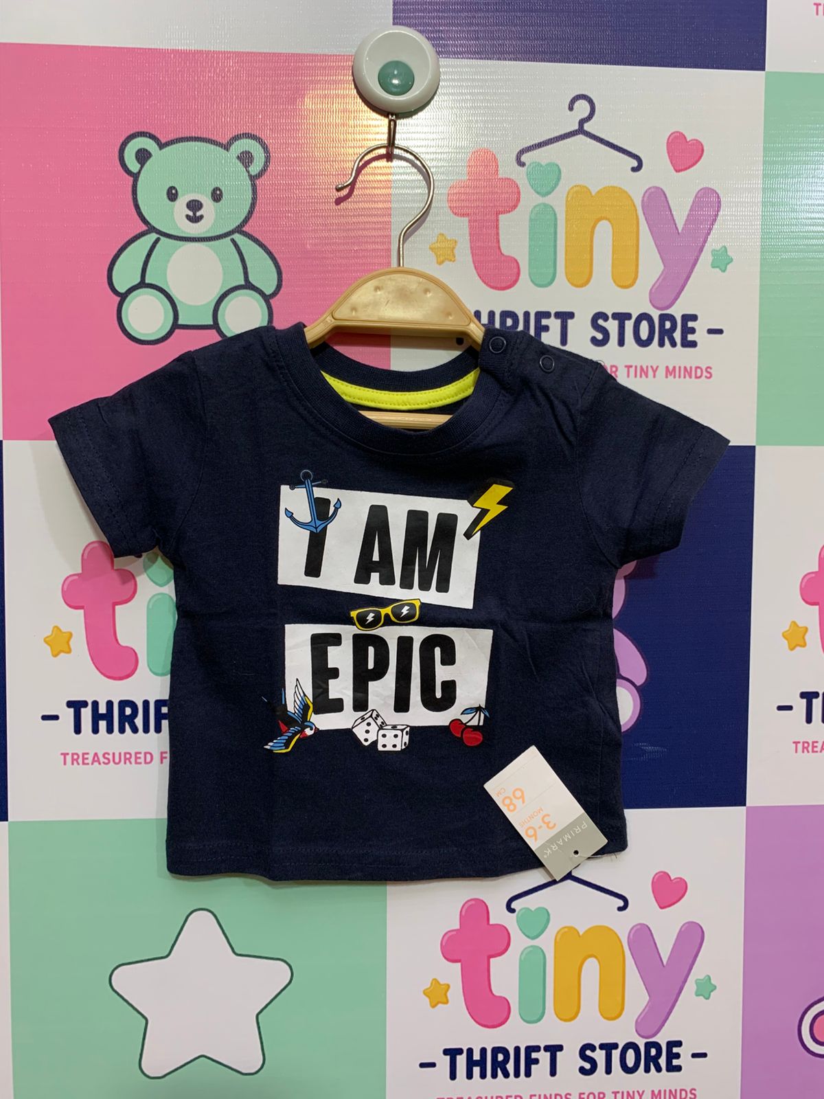 Front view of a dark navy baby t-shirt with "I AM EPIC" printed in bold white blocks, surrounded by small icons like a yellow lightning bolt and red cherries. Close-up shows the Primark brand label and size tag "9-12 Months / 80cm". Background is a clean studio-style white.