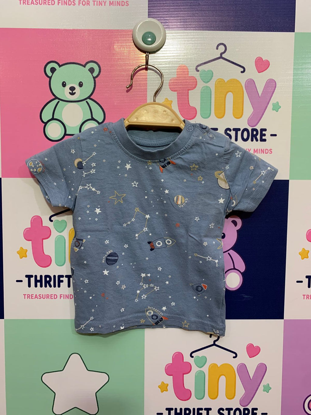 A light blue baby t-shirt with a detailed all-over print of rockets, stars, and planets. Close-up shows the Primark brand label inside the neck with size "0-3 Months / 62cm". Background is a clean studio-style white.