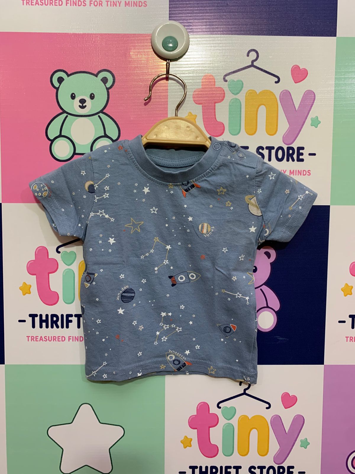 A light blue baby t-shirt with a detailed all-over print of rockets, stars, and planets. Close-up shows the Primark brand label inside the neck with size "0-3 Months / 62cm". Background is a clean studio-style white.