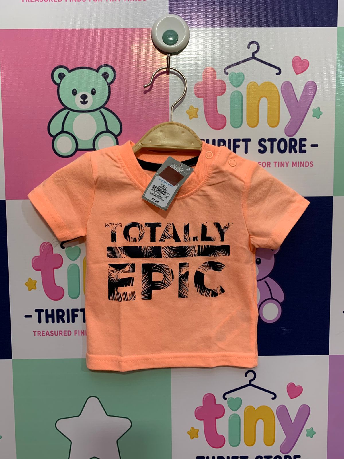 Front and back view of a small neon peach infant t-shirt by Primark hanging on a hanger. It features a bold black text print on the chest reading "TOTALLY EPIC". Close-up shows the brand label "PRIMARK" and the size/care label reading "SIZE 0–3 Months / 62CM" and "MADE IN INDIA". The backdrop is a colorful thrift store checkered wall.