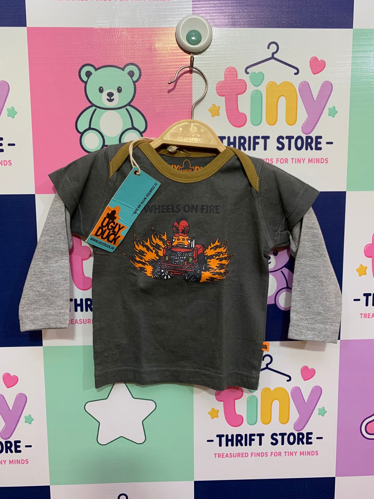 Front and back view of a small grey double-layer infant t-shirt by Ugly Duck hanging on a hanger. It features contrasting heather grey sleeves with a bold monster truck graphic on the chest reading "WHEELS ON FIRE". Close-up shows the brand label "UGLY DUCK" and the size stamp label reading "SIZE 4-6M / 68cm". The backdrop is a colorful thrift store checkered wall.