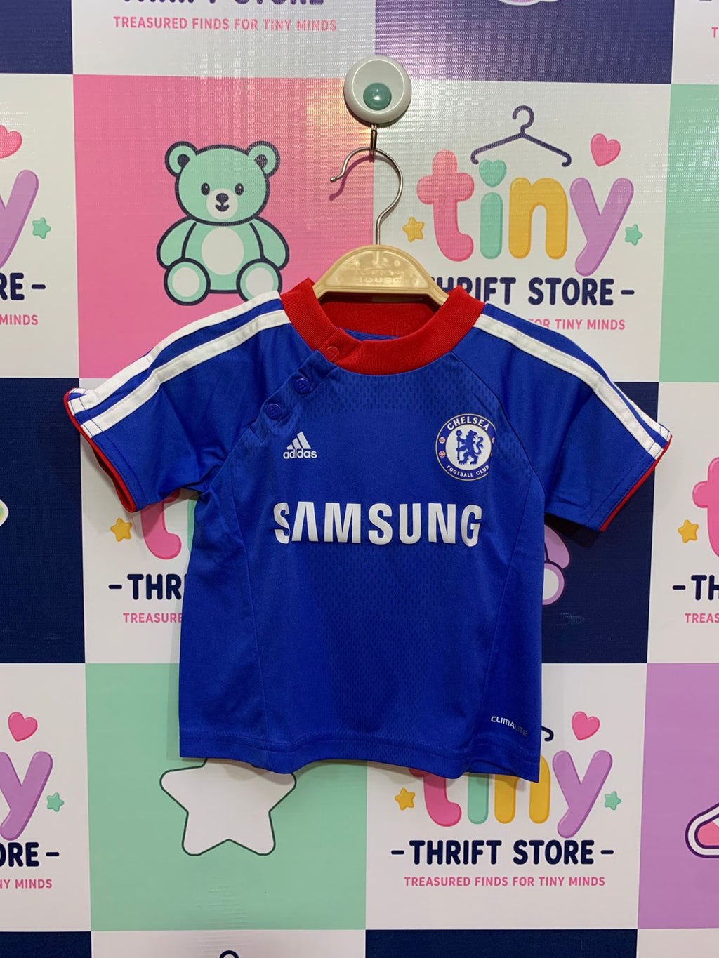 Front and back view of a small royal blue infant football jersey for Chelsea FC, hanging on a hanger. It features the Adidas logo, official club crest, and a bold white 'SAMSUNG' sponsor logo on the chest. The collar is a contrasting red ribbed fabric. A small white text tag on the collar placket specifies "CLIMALITE" fabric technology. A close-up shows the brand label "ADIDAS" and the size label reading "SIZE 3-6 MONTHS / 62CM" and "MADE IN INDIA". The backdrop is a colorful thrift store checkered wall.