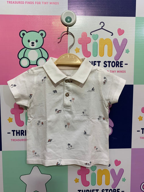 Front and back view of a small white infant t-shirt by C&A hanging on a wooden hanger. It features a charming nautical pattern of tiny sailboats, palm trees, and huts all over, with contrasting brown buttons. Close-up shows a soft inner collar stamp label reading "C&A Newborn / Size 74 6-9M" and "designed with love". The backdrop is a colorful thrift store checkered wall.