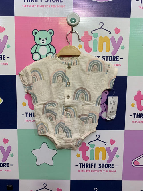 A close-up view of a brand new cream cotton muslin baby romper from George with original tags attached. The romper features an all-over print of colorful pastel rainbows and two small brown buttons. The attached tag displays the price £6.50. Another close-up photo shows the fabric labels confirming the brand "GEORGE CLOTHING" and size "0–3 MONTHS" up to 12lb/5.5kg, and "made with better cotton". The romper hangs on a hanger.