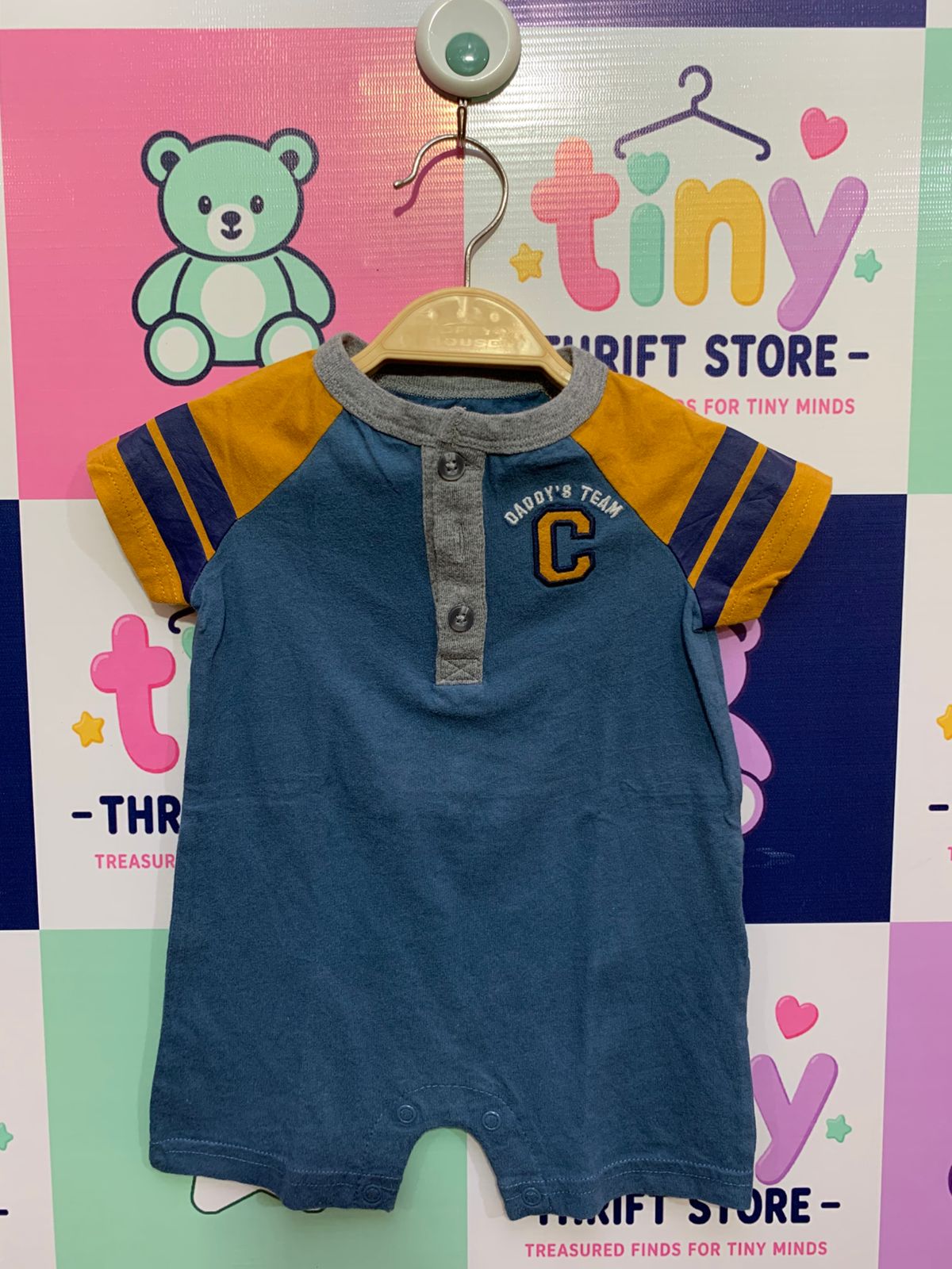 Front and back view of a small dusty blue and mustard yellow infant romper by Carter's hanging on a hanger. It features striped sleeves, a grey-ribbed collar placket with buttons, and an embroidered chest motif reading "DADDY'S TEAM" and a large letter "C". A close-up shows the brand label "CARTER'S" and the size label reading "SIZE 6M / MADE IN CAMBODIA". The backdrop is a colorful thrift store checkered wall.
