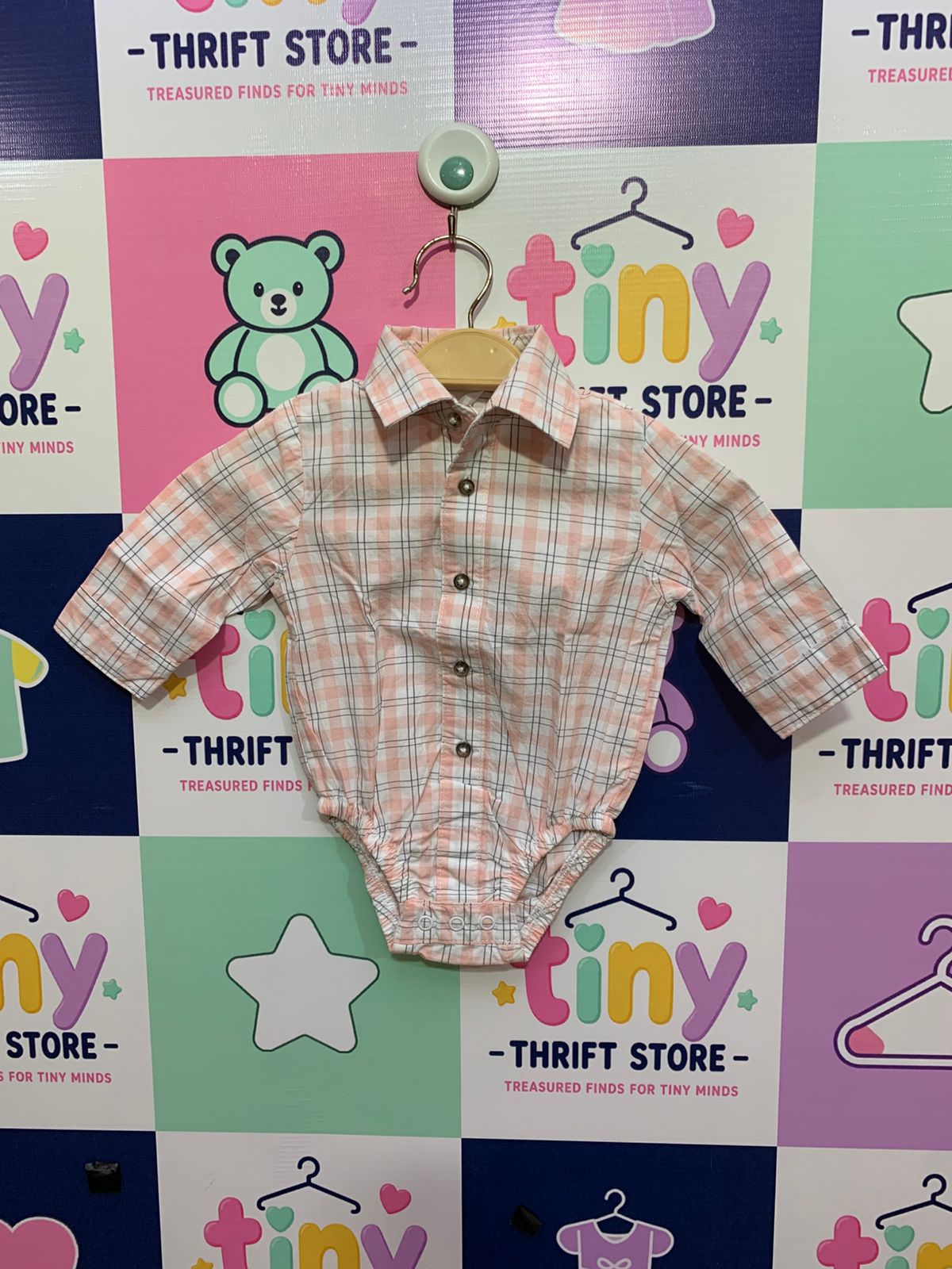 A baby boy's peach and grey plaid long-sleeve button-down bodysuit displayed on a hanger against a colorful "Tiny Thrift Store" backdrop.