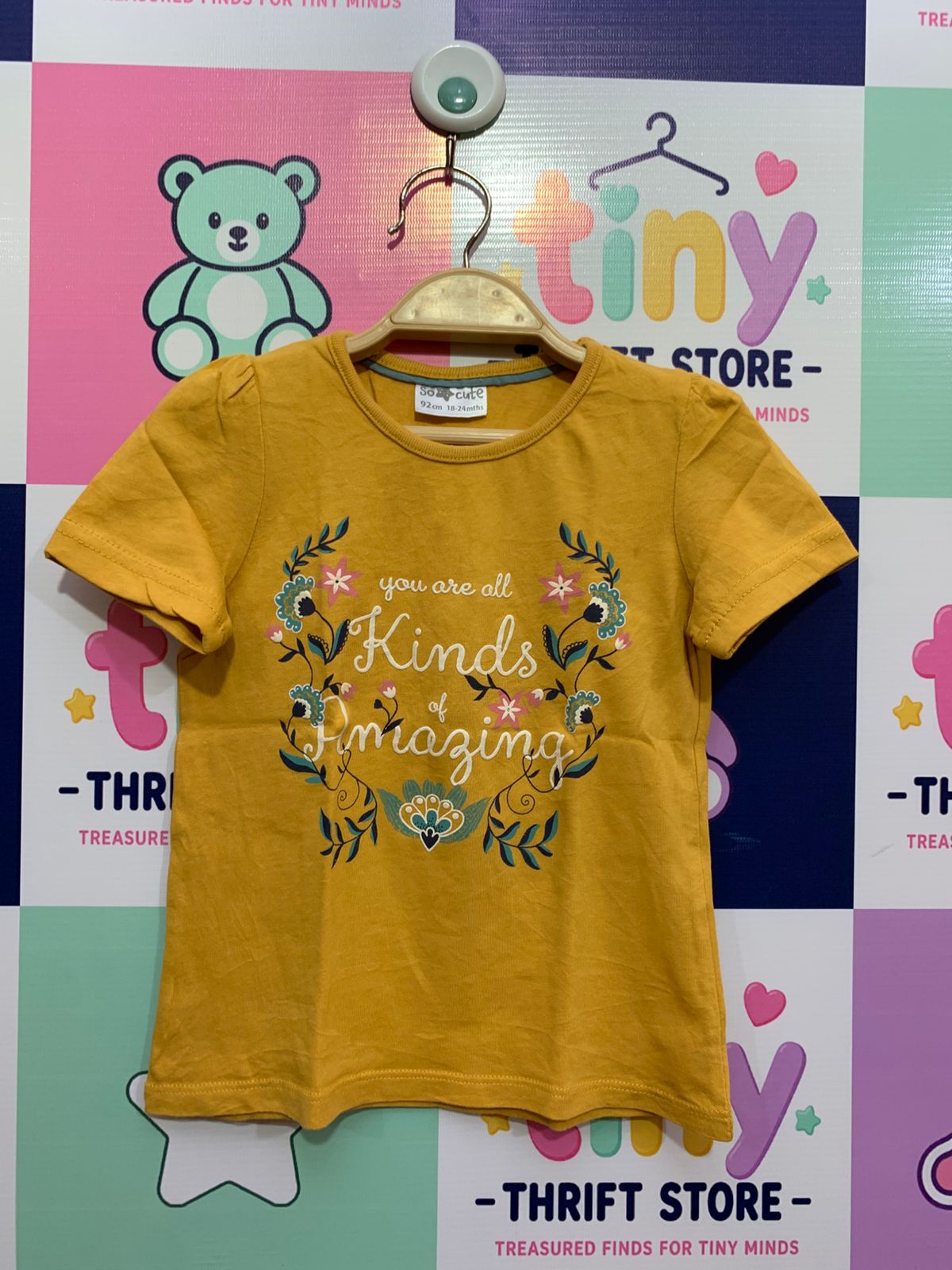 Front and back view of a "So Cute" mustard yellow toddler t-shirt with floral graphics and "You are all kinds of Amazing" text, displayed on a Tiny Thrift Store background
