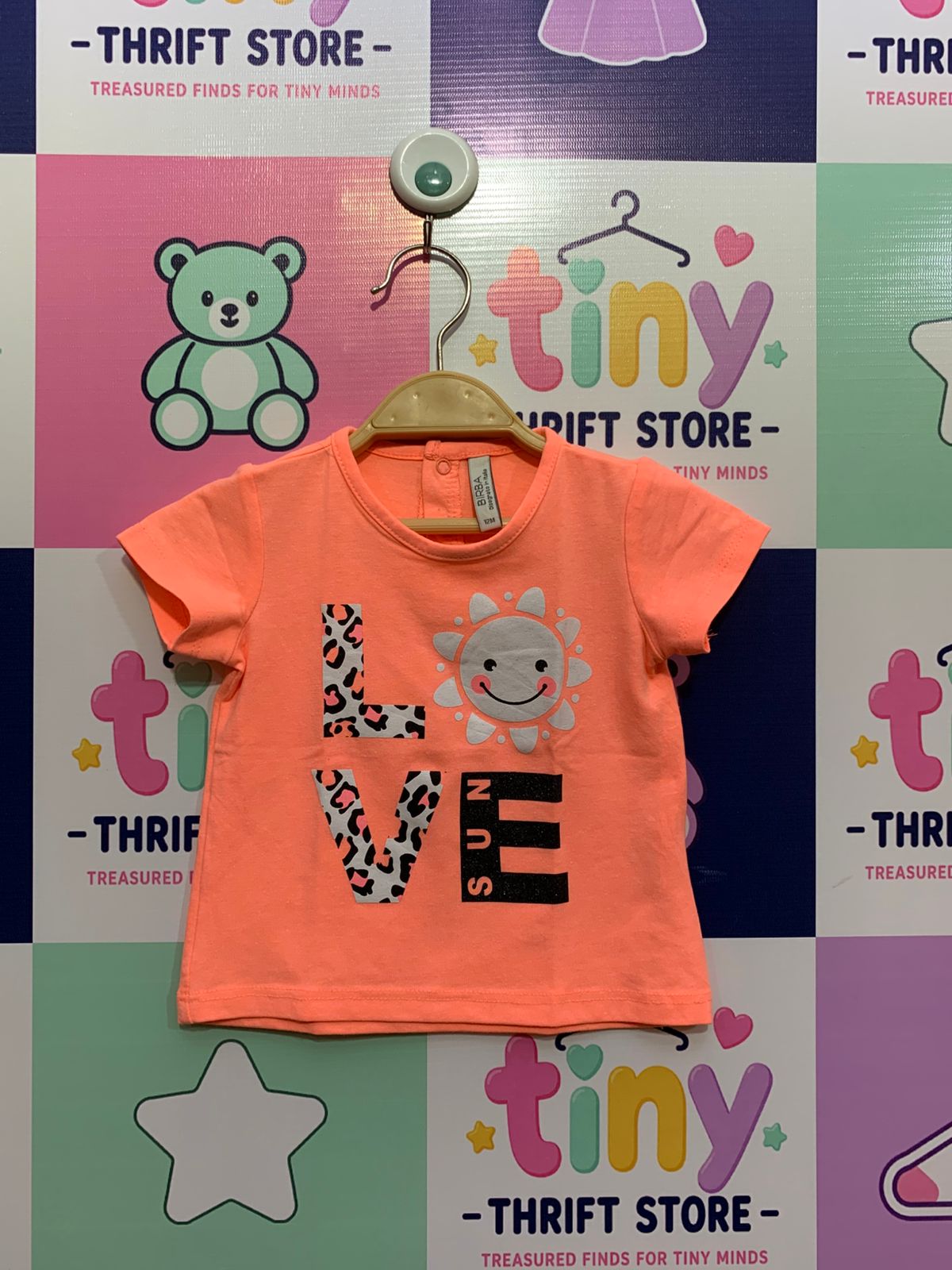 Front and back view of a Birba neon peach baby t-shirt featuring "LOVE" text in leopard print and a sun graphic, hanging on a Tiny Thrift Store background