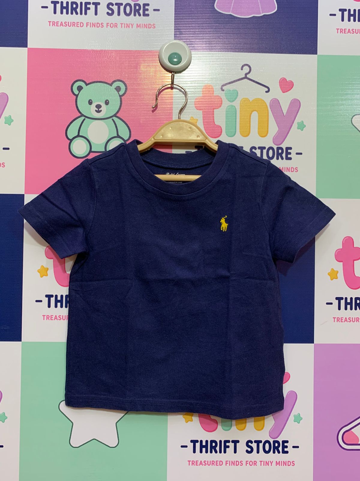 Front and back view of a Ralph Lauren navy blue baby t-shirt for 18 months, featuring a yellow polo player logo, hanging on a Tiny Thrift Store backdrop