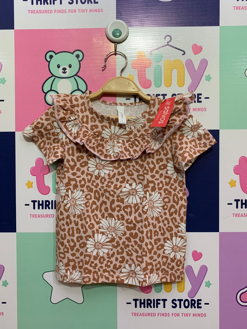 A baby girl's soft, short-sleeve Dymples top featuring an all-over leopard print mixed with white daisies. It has a distinctive layered ruffle collar with subtle pink stitching, hanging on a hanger against a colorful "Tiny Thrift Store" backdrop