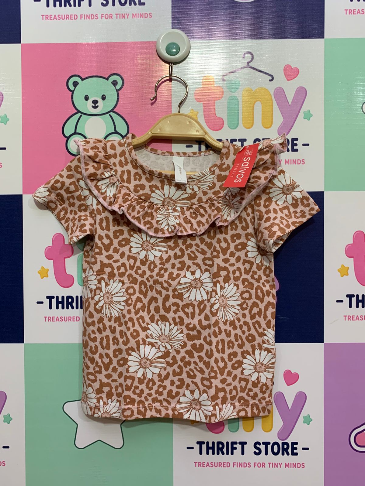 A baby girl's soft, short-sleeve Dymples top featuring an all-over leopard print mixed with white daisies. It has a distinctive layered ruffle collar with subtle pink stitching, hanging on a hanger against a colorful "Tiny Thrift Store" backdrop