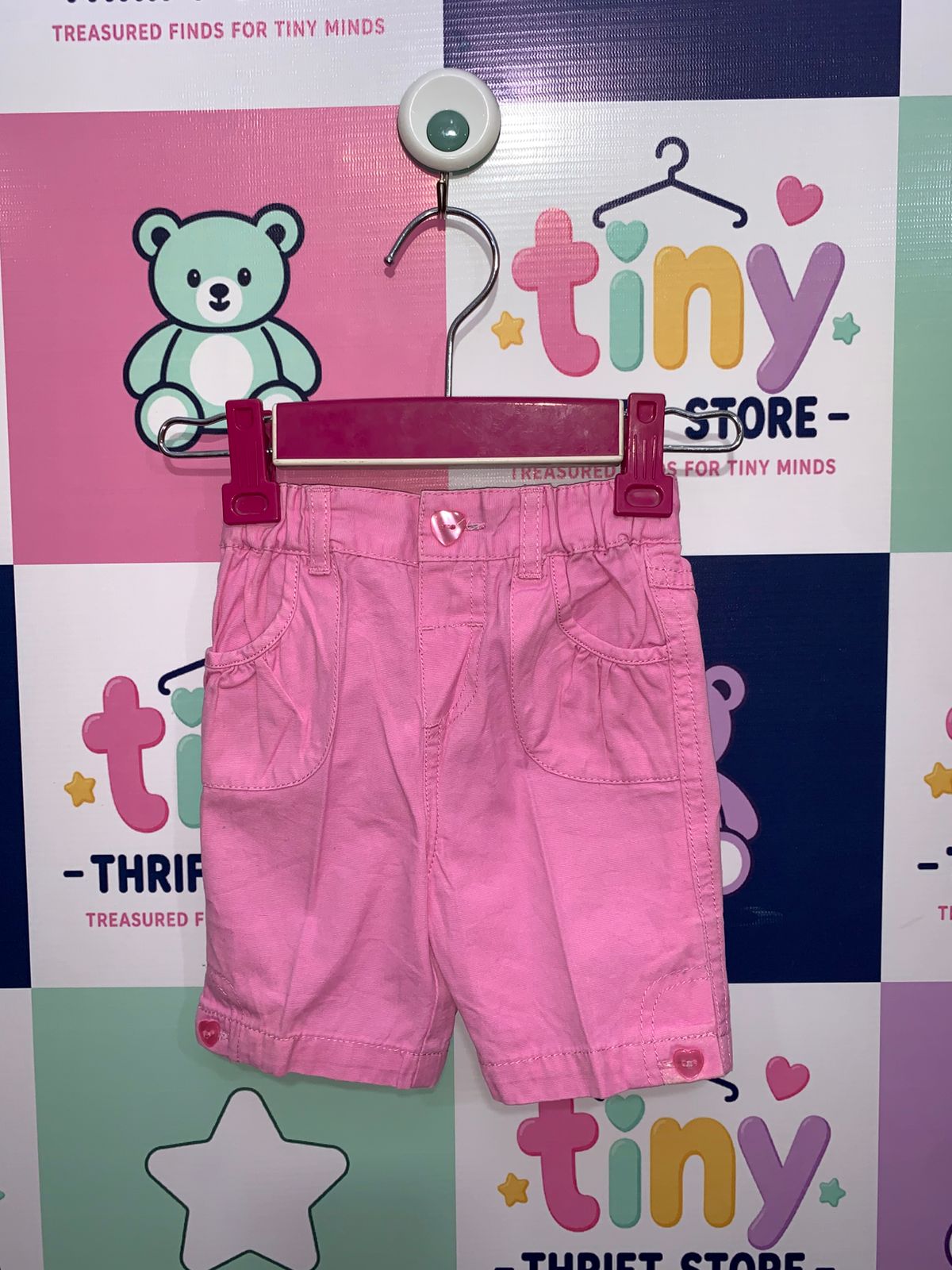 A pair of bright pink baby girl shorts from Early Days brand, size 6-9 months, featuring an elastic waistband, heart-shaped buttons, and front pockets, displayed on a hanger against a Tiny Thrift Store background.