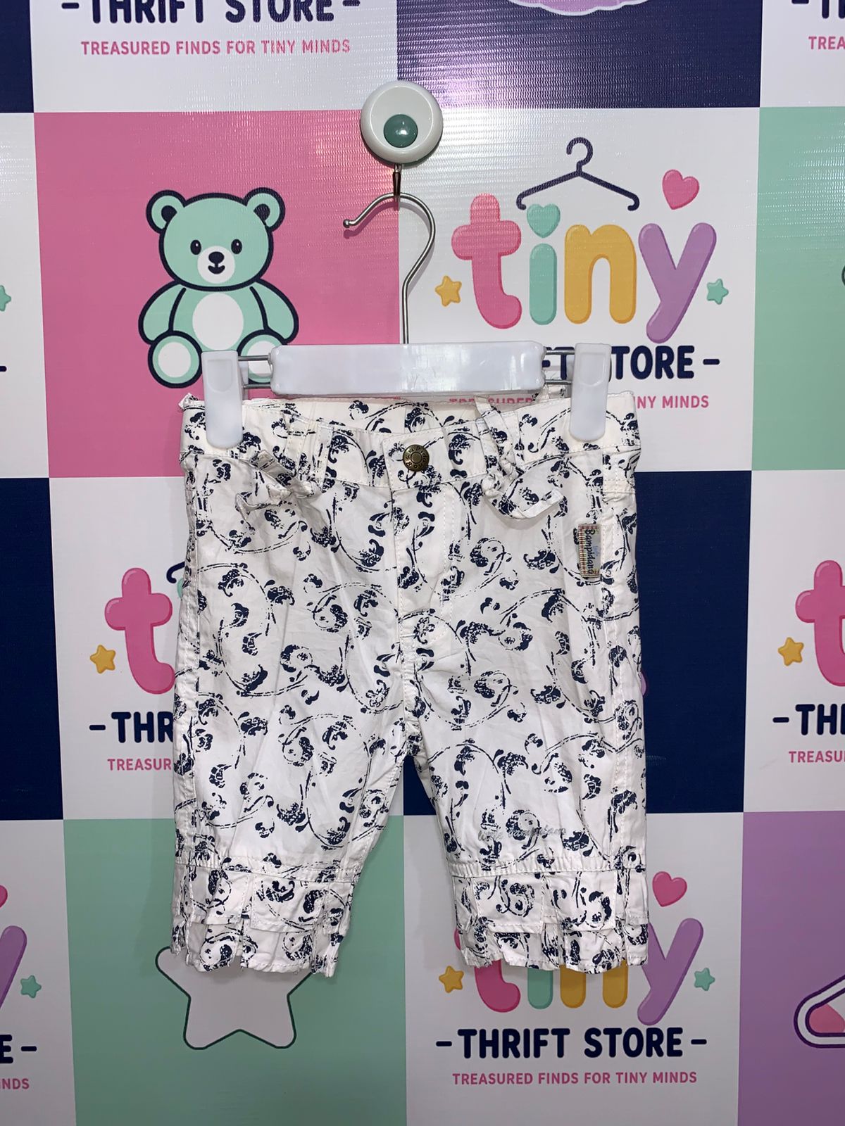 A pair of white girls' capri pants with a navy blue floral print hanging on a white hanger against a colorful Tiny Thrift Store backdrop. The pants feature ruffle hems and small bows at the waist.