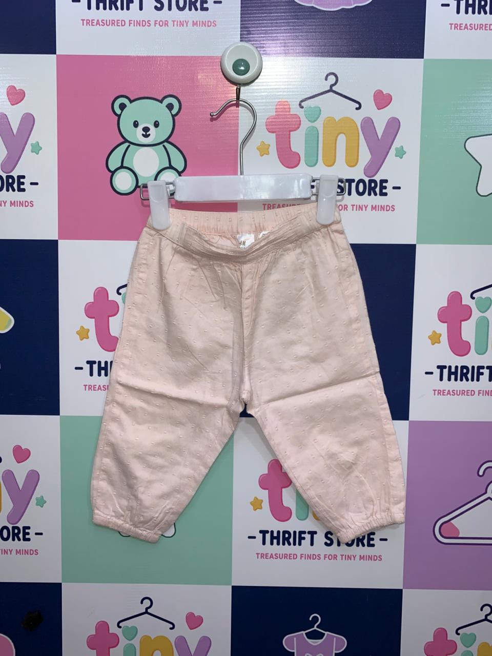 A front view of H&M baby girl peach trousers featuring a textured dobby dot pattern, hanging on a white hanger against a "Tiny Thrift Store" branded background. The trousers have an elasticated waistband and gathered hems at the ankles.