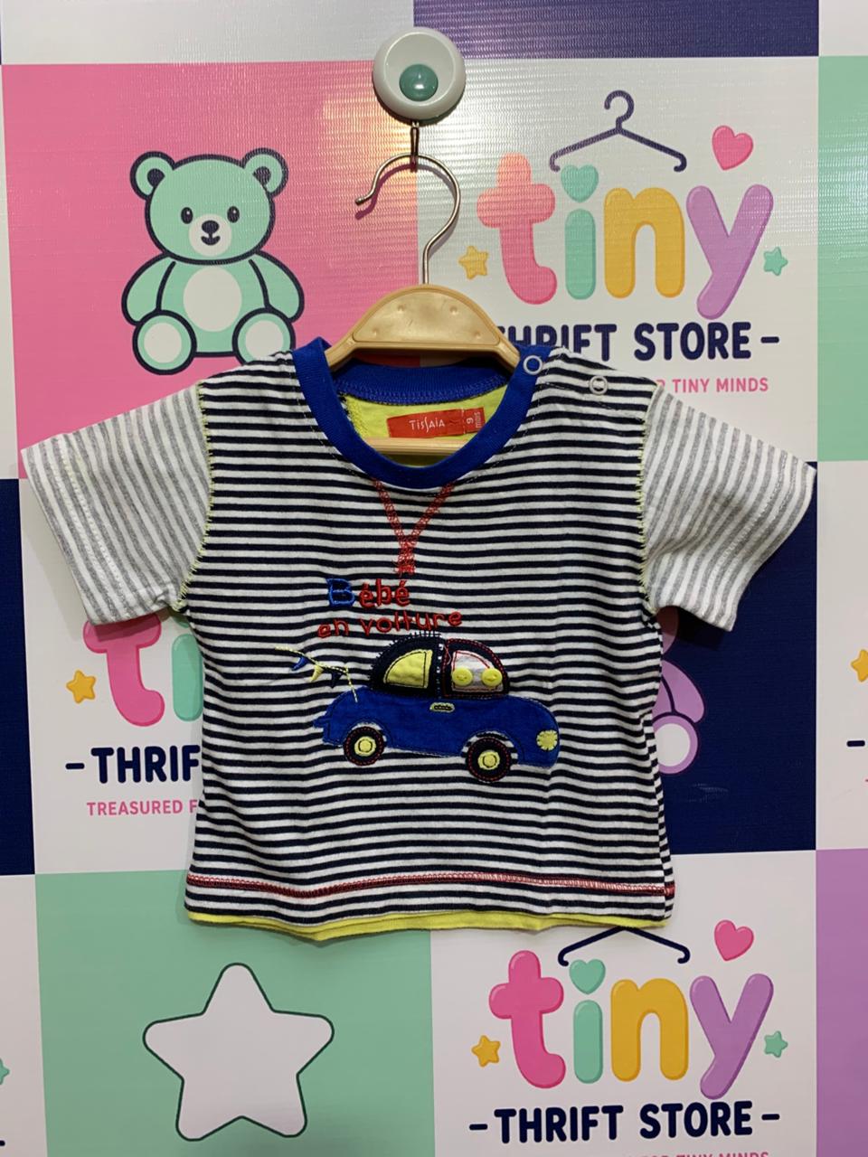 Front and back view of a small baby boy's t-shirt hanging on a wooden hanger. It features alternating navy-and-white and grey-and-white stripes with a large embroidered blue car on the chest. The French words "Bebé en voiture" are stitched in blue and red. The inside collar shows a red label with the brand "TISSAIA" and the size "9m". The backdrop is a colorful checkered thrift store wall.